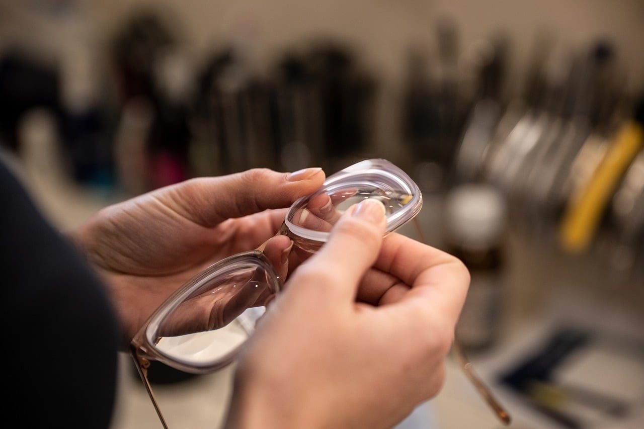 An optician's hands holding a spectacle lens up for inspection at a workbench.