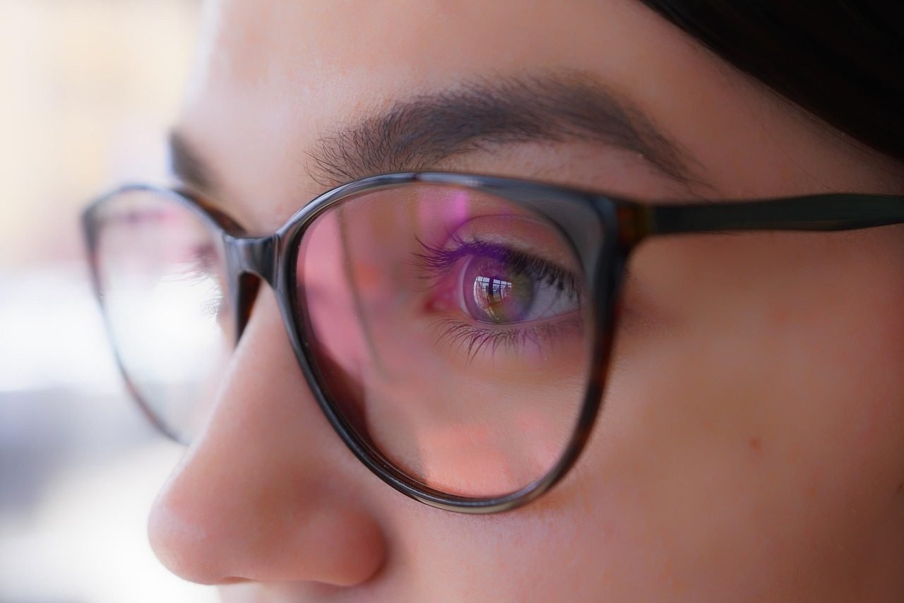 Close-up of an eye behind a spectacle lens, with a violet flash from the anti-reflective coating.